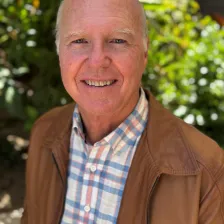 Don Mitchell smiling outdoors, wearing a brown jacket over a plaid shirt, stands in front of lush green foliage. The mood is warm and cheerful.