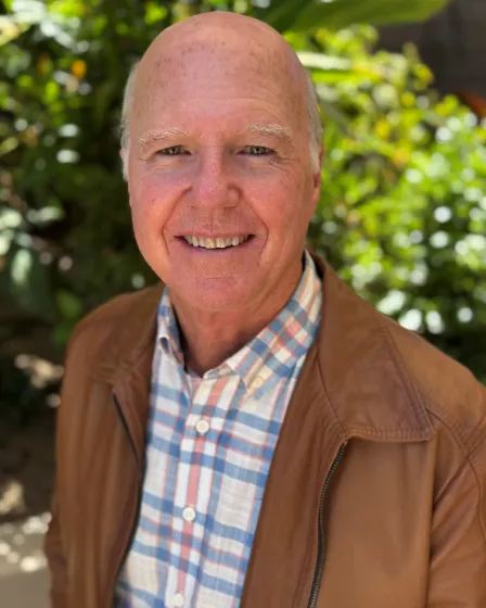 Don Mitchell smiling outdoors, wearing a brown jacket over a plaid shirt, stands in front of lush green foliage. The mood is warm and cheerful.